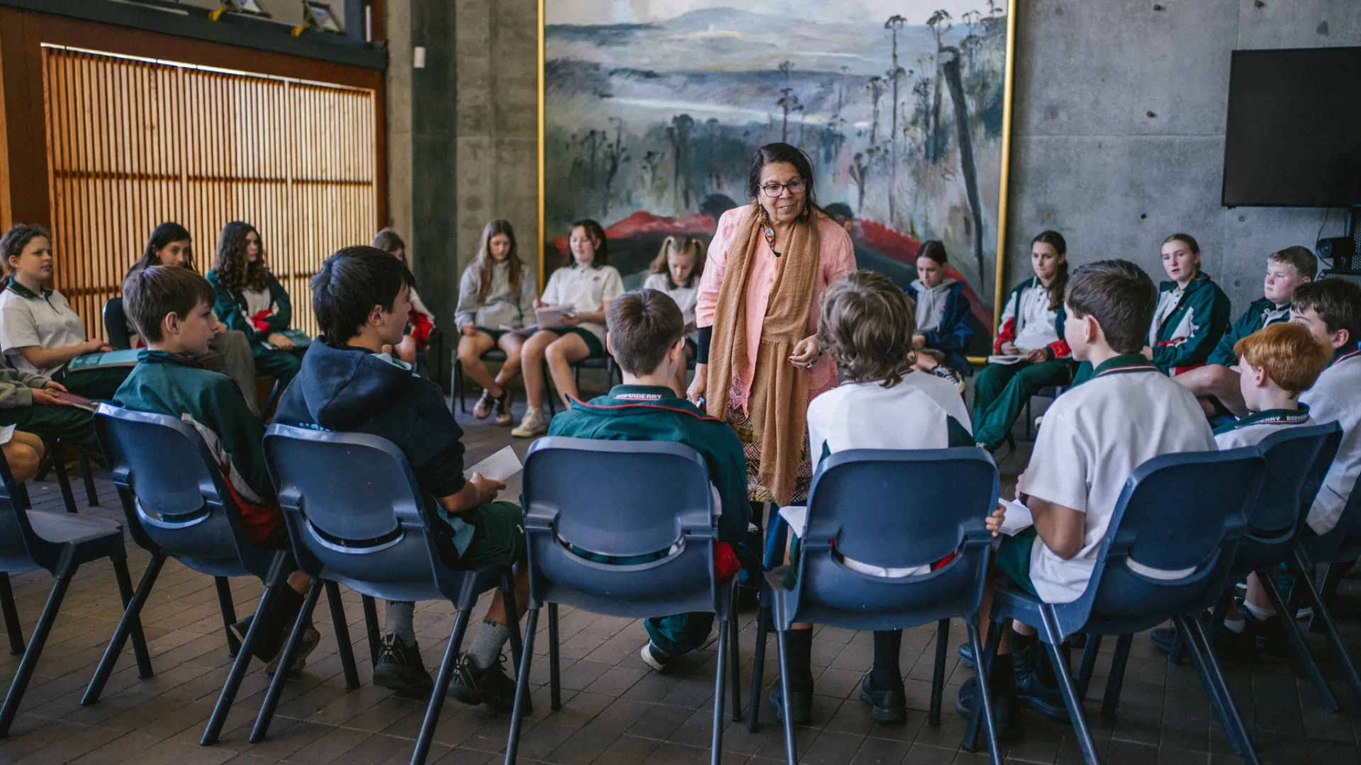 A woman stands, speaking in the middle of a circle of primary school students sitting in plastic blue chairs in a circle. They are in a large room, that has a floor to ceiling nature painting behind them.