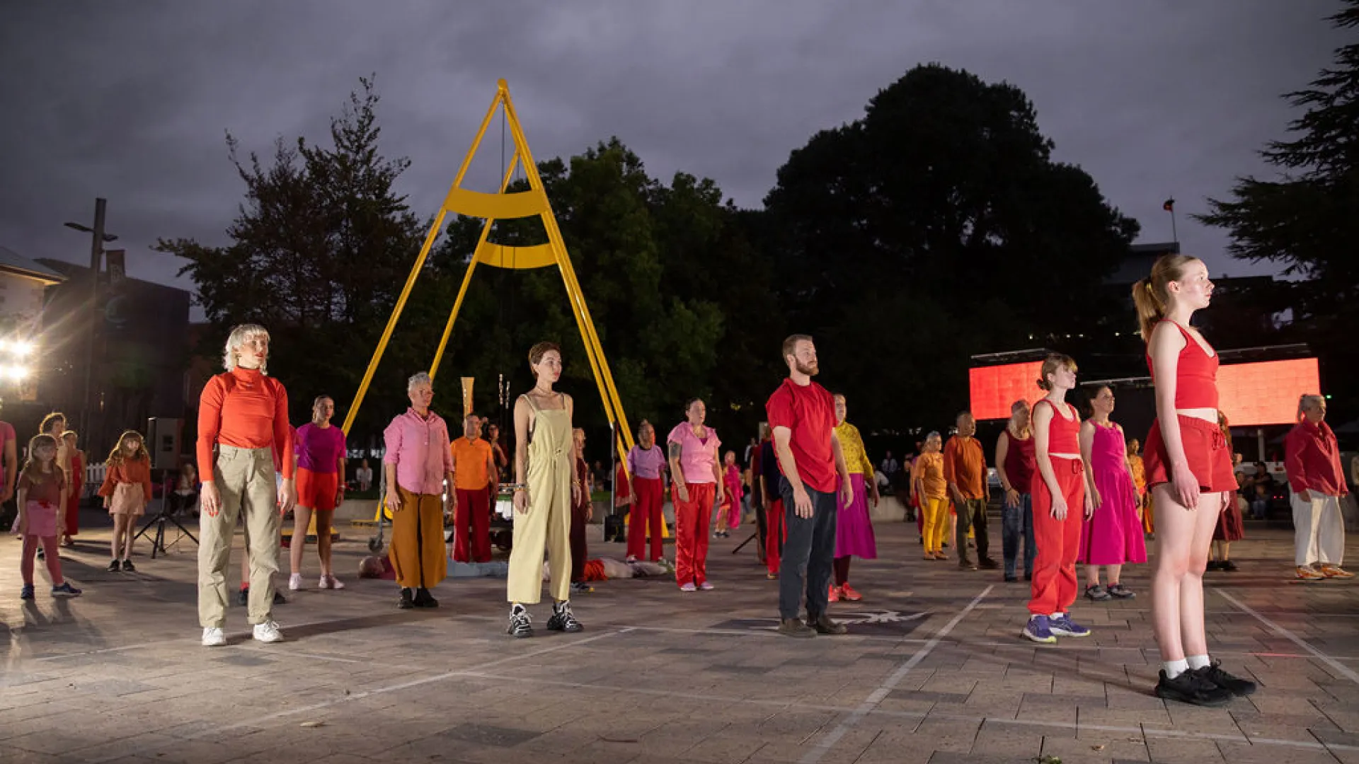 A photo of dancers all standing straight, tall and not moving. They are standing in a carpark, spread out but all facing the same way. They wear colourful clothing, but the image is eerie.